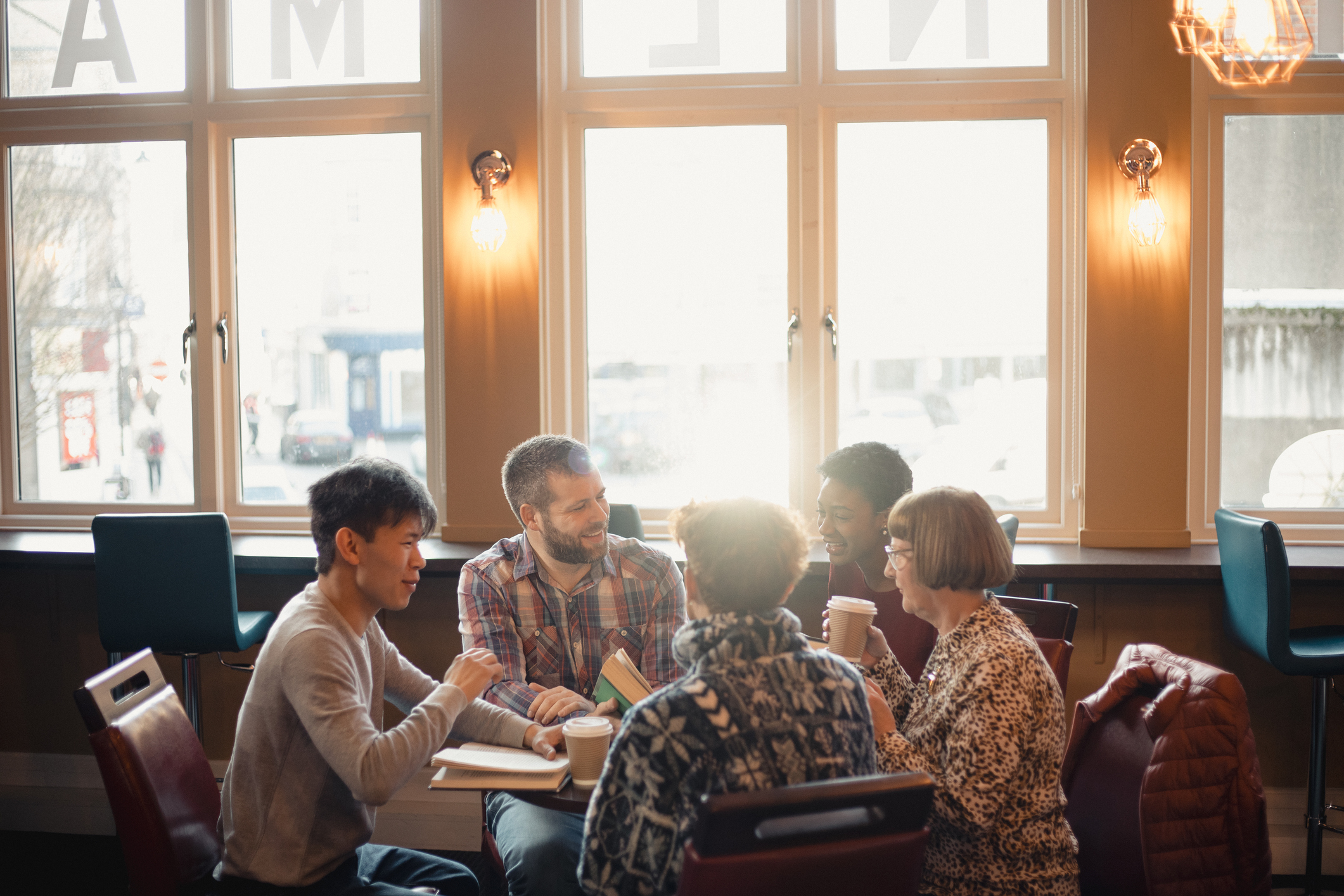 Small group of people with a mixed age range sitting around a table in a cafe. They are talking during a book club.