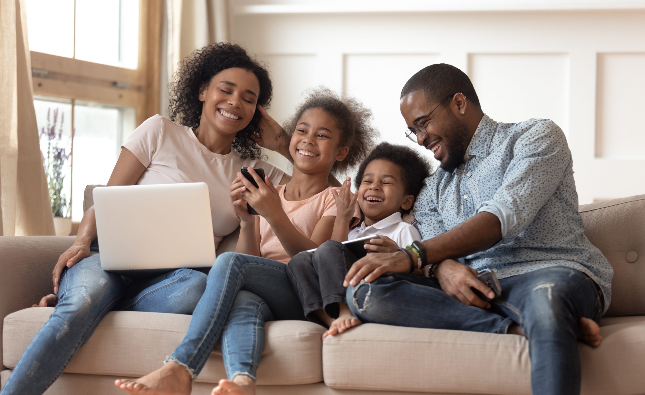 Cheerful african parents and kids laugh use devices together sit on sofa, tech addicted family with children hold laptop phone digital tablet having fun with gadgets at home, technology dependence